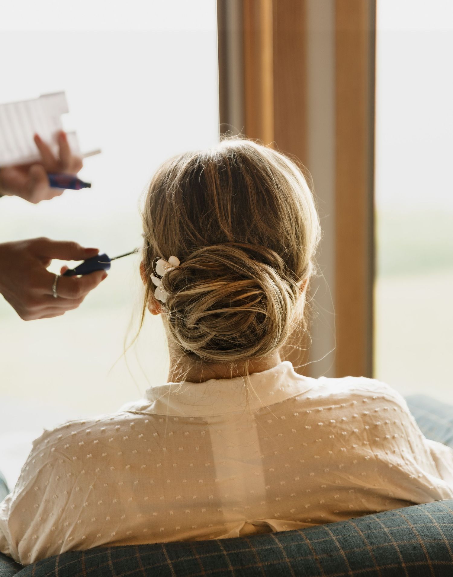 A hairstylist finishing a elegant low bun updo on a blonde woman near a bright window.