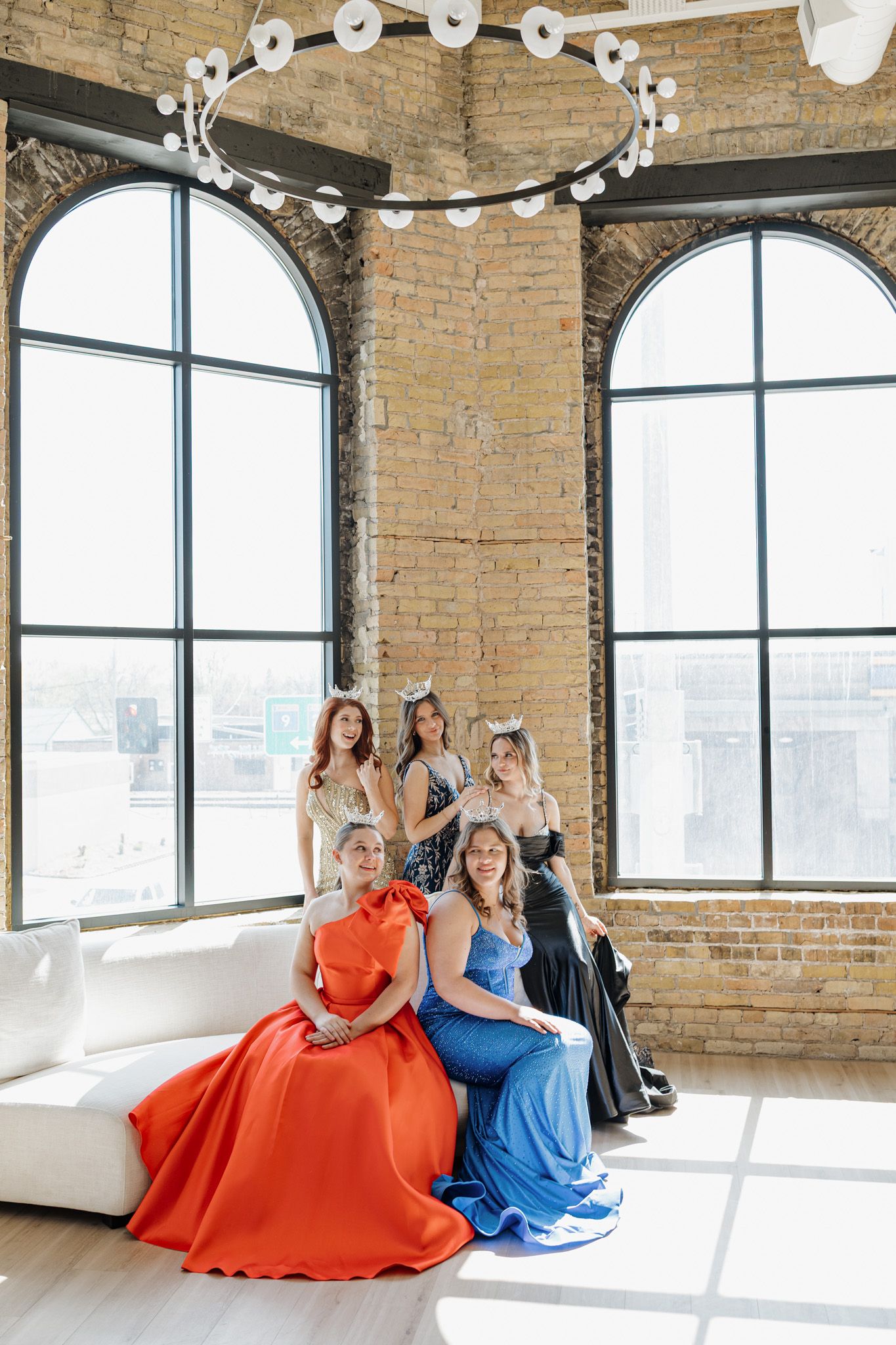 Five pageant contestants in elegant gowns and crowns pose in a sunlit brick loft studio.