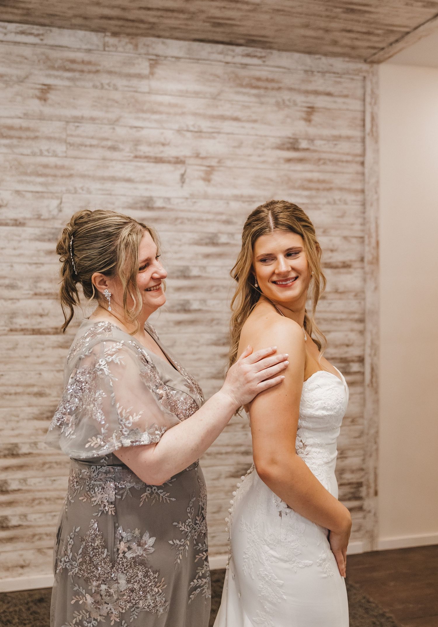 Bride in white lace gown smiles as woman in floral gray dress helps her prepare before the wedding ceremony.