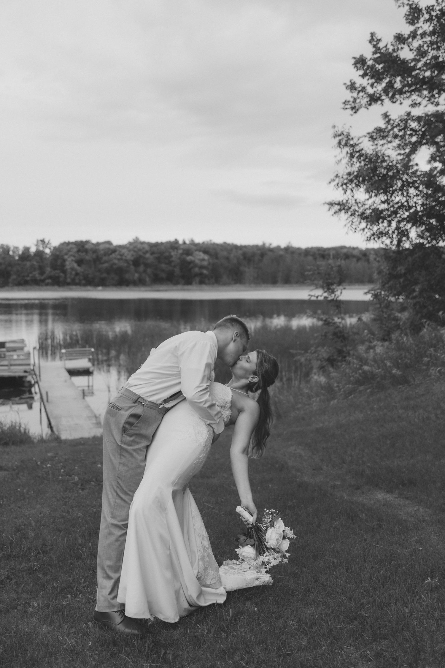 Black and white photo of a bride and groom sharing a romantic dip kiss by a serene lake on their wedding day.