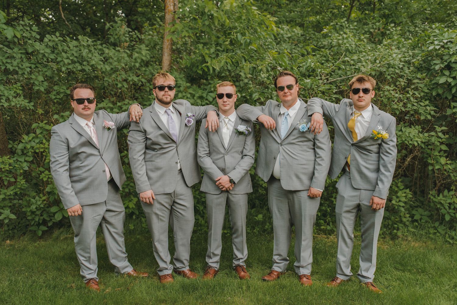 Five groomsmen in matching gray suits and sunglasses pose outdoors on a green lawn at a wedding.