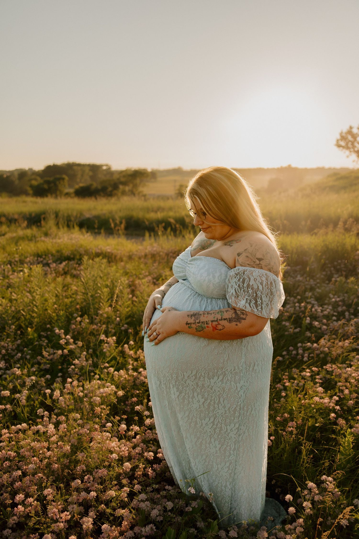 Pregnant woman in light blue lace gown cradling her belly in a sunlit wildflower field at golden hour.