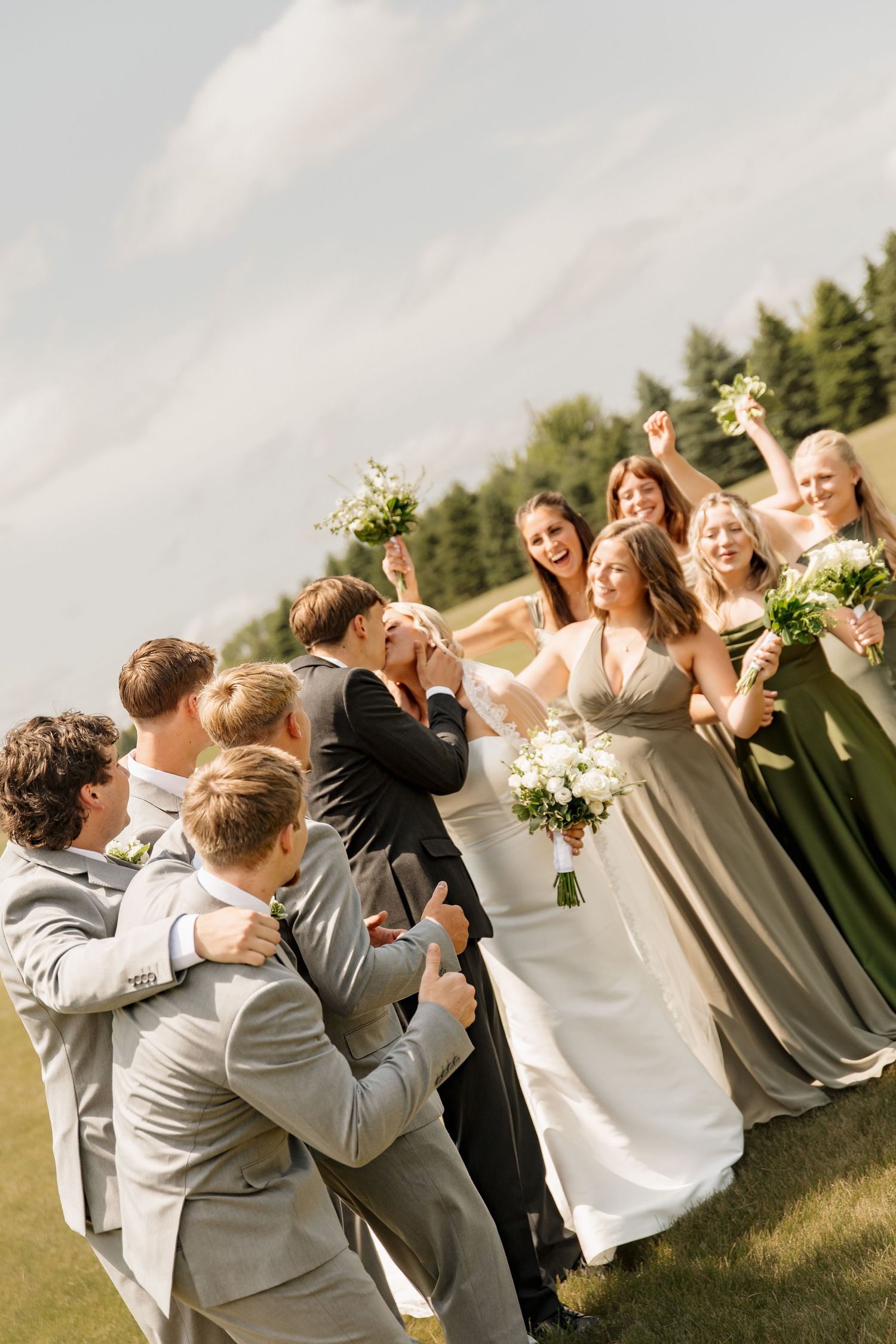 Joyful wedding party celebrating on green lawn with bridesmaids in sage gowns and groomsmen in gray.