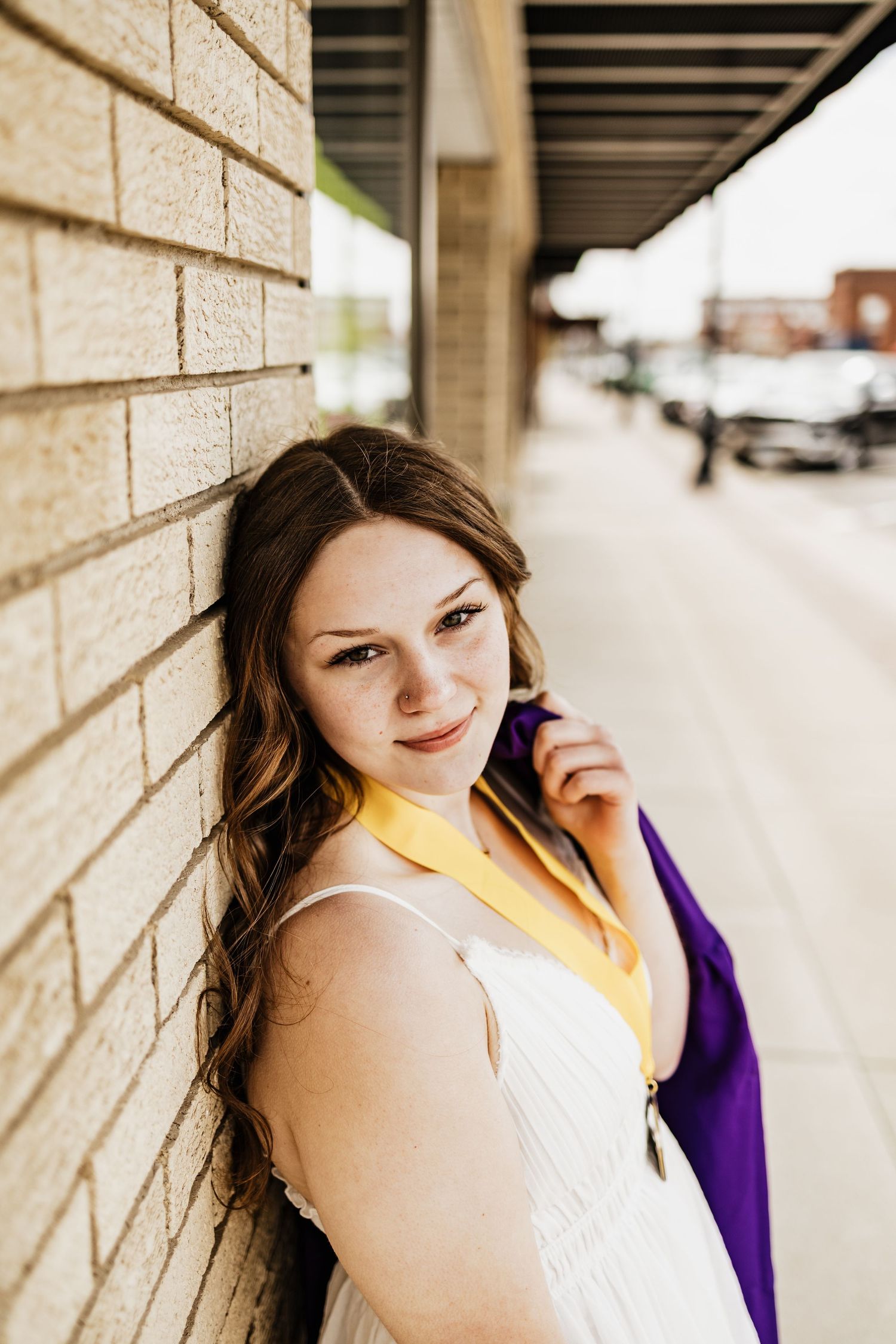 Young woman in white dress with purple and gold graduation regalia leaning against brick wall outdoors.