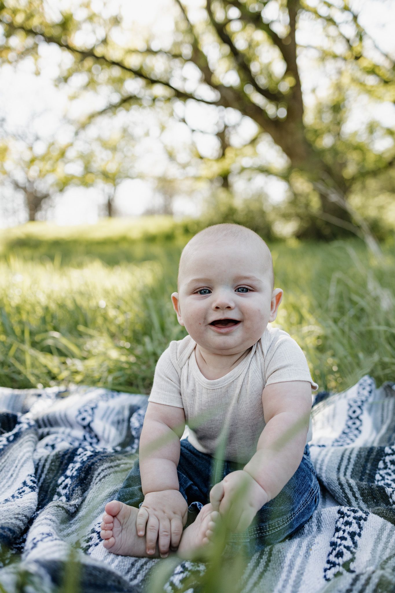 Happy baby sitting on a blanket outdoors in green grass, smiling at camera with a large tree in the background.