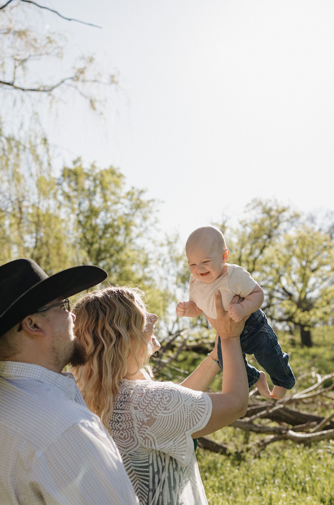 A couple lifts their smiling baby in the air outdoors on a sunny day surrounded by green trees.