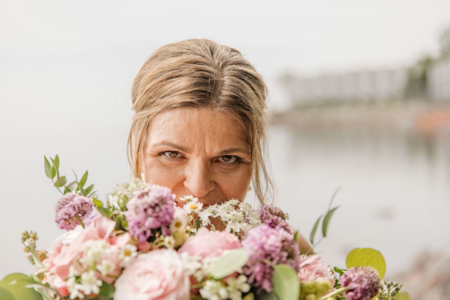 A bride holds a lush pink and purple bouquet up to her face near a waterfront on her wedding day.