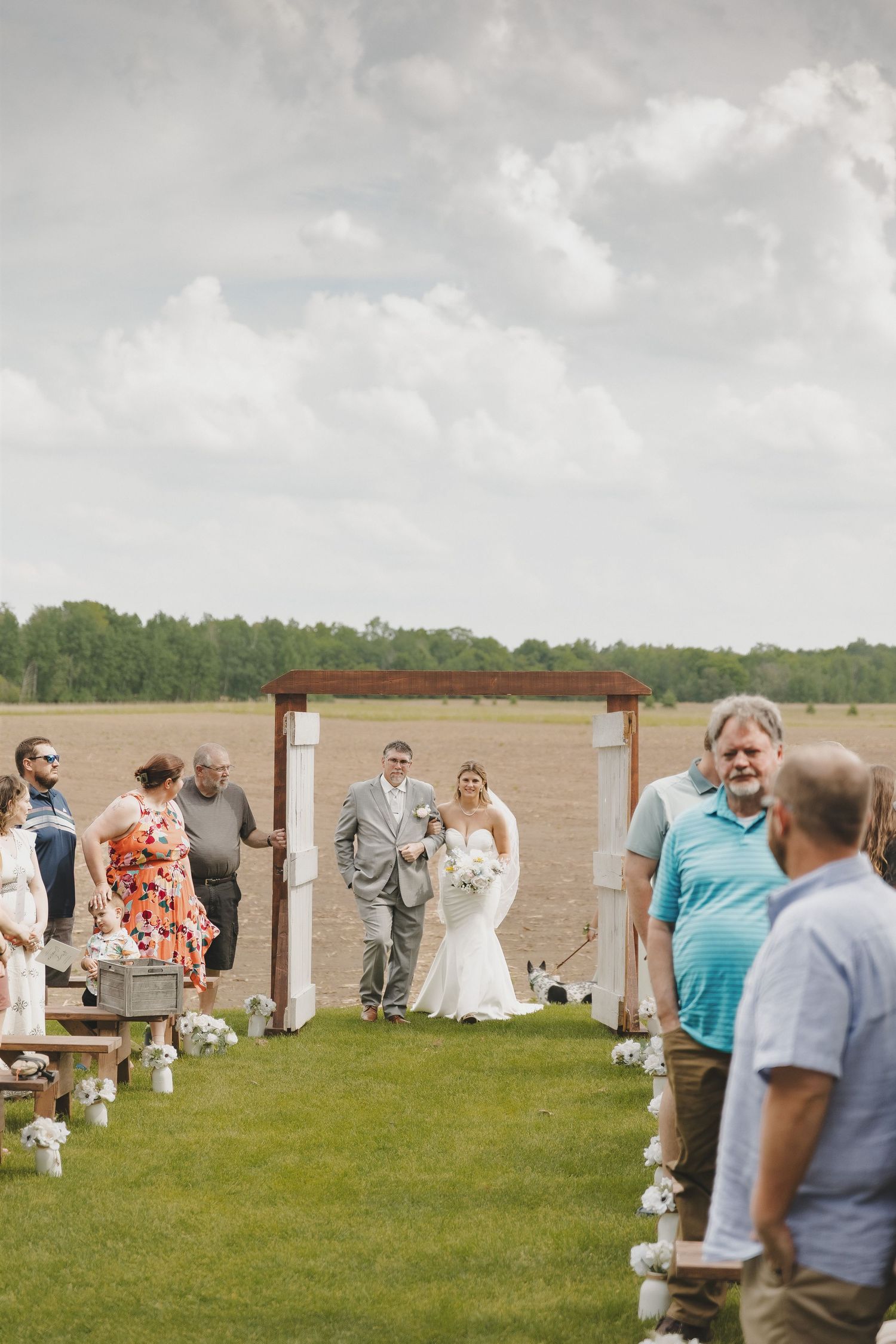 Bride walks down outdoor aisle with escort at rustic farm wedding ceremony under cloudy sky.