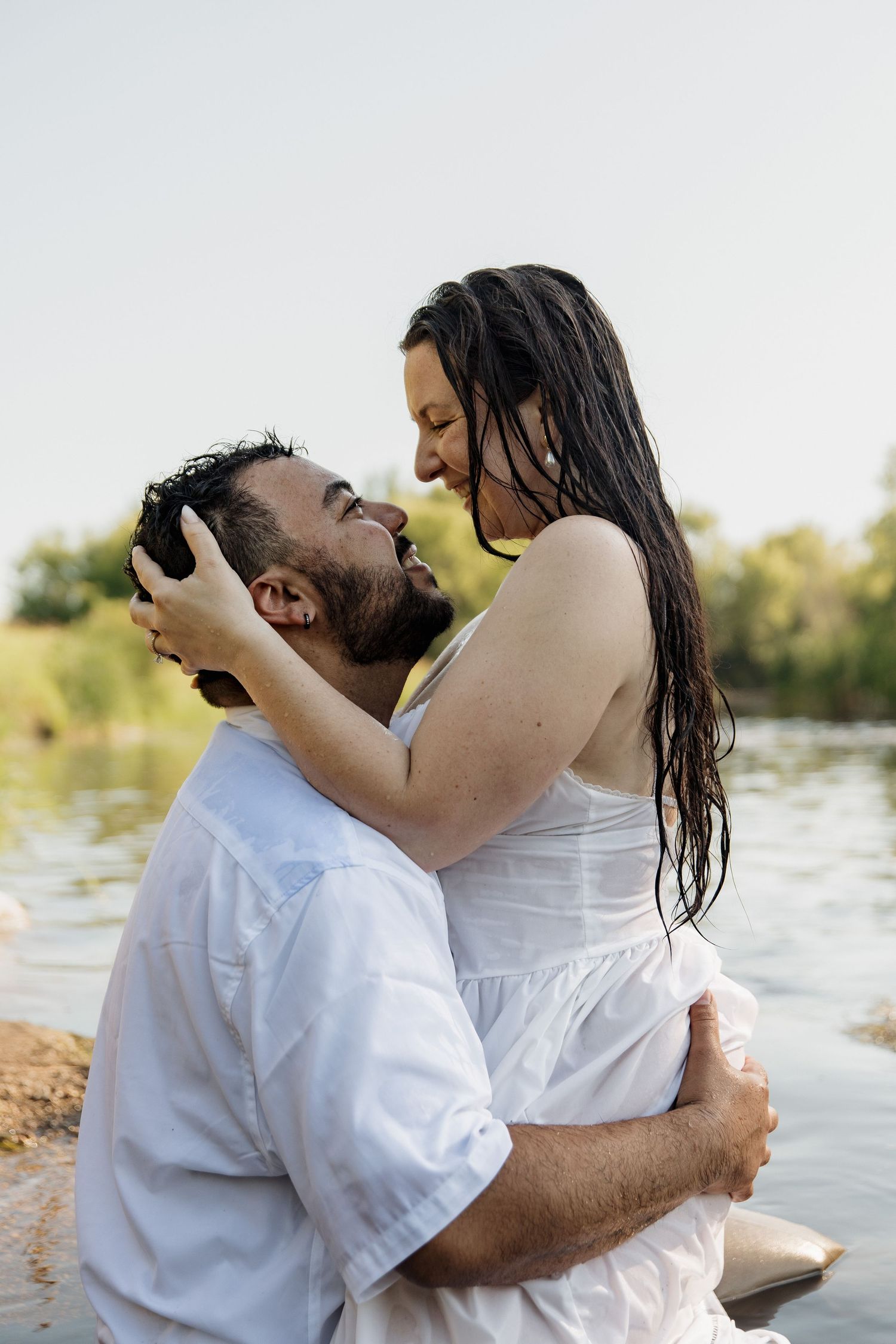 Romantic couple in white embracing by a river, woman with wet hair holding man's face as they gaze at each other.