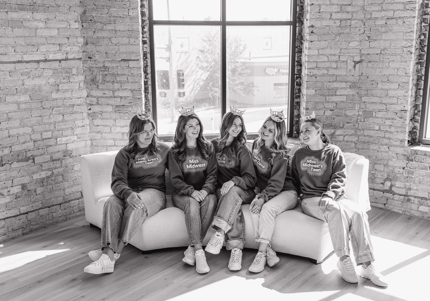 Five women in matching sweatshirts sit together laughing on white sofa in brick-walled loft space.