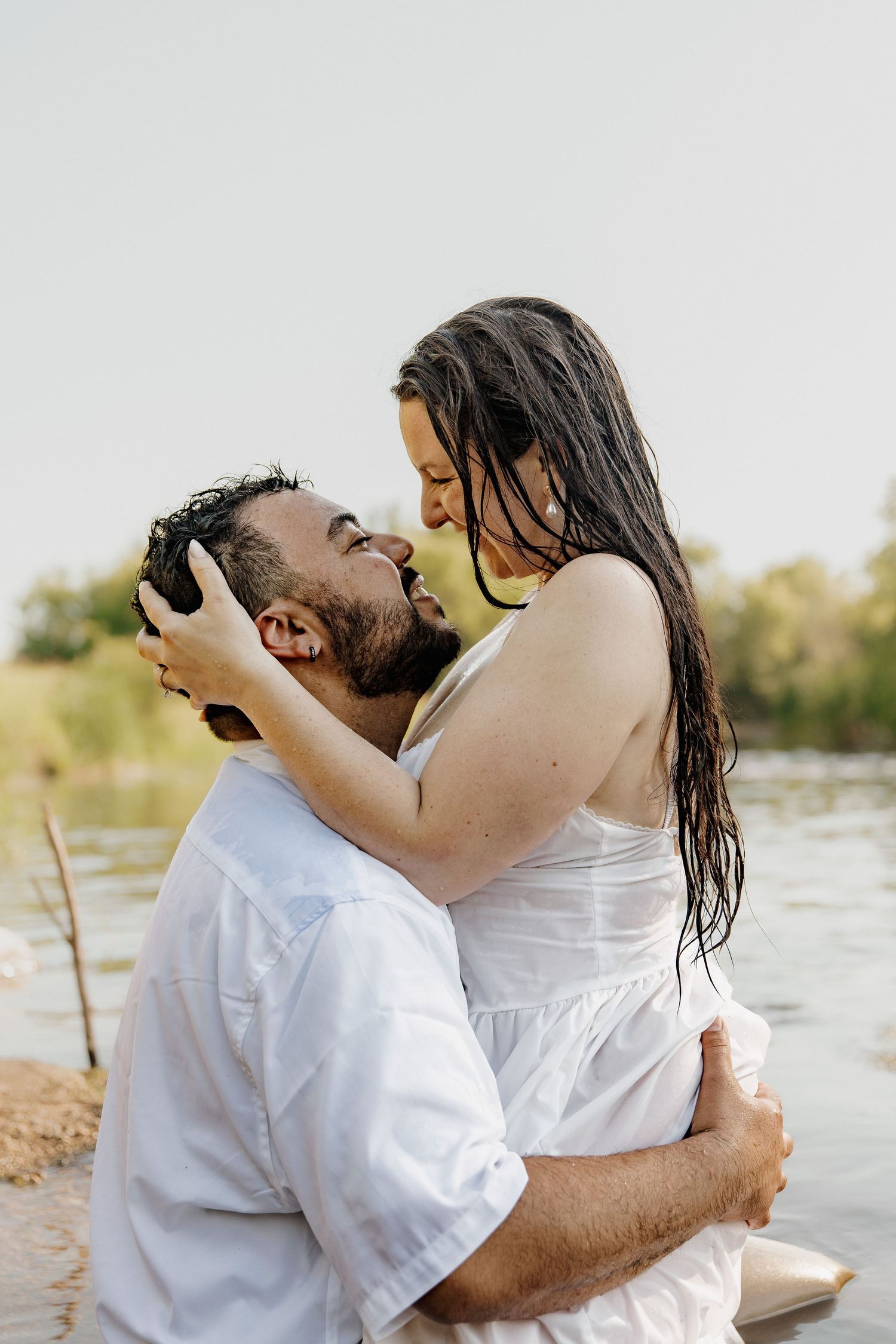 Couple in white clothing embracing near a river, woman with wet hair holding man's face as they share a romantic moment.