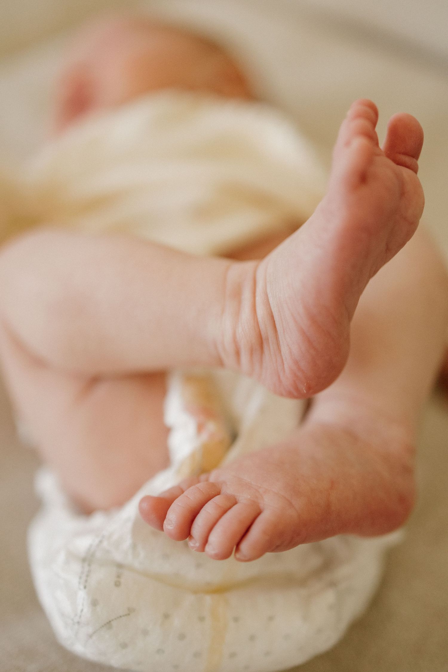 Close-up of tiny newborn baby feet and toes, with infant lying on white diaper in soft natural light.