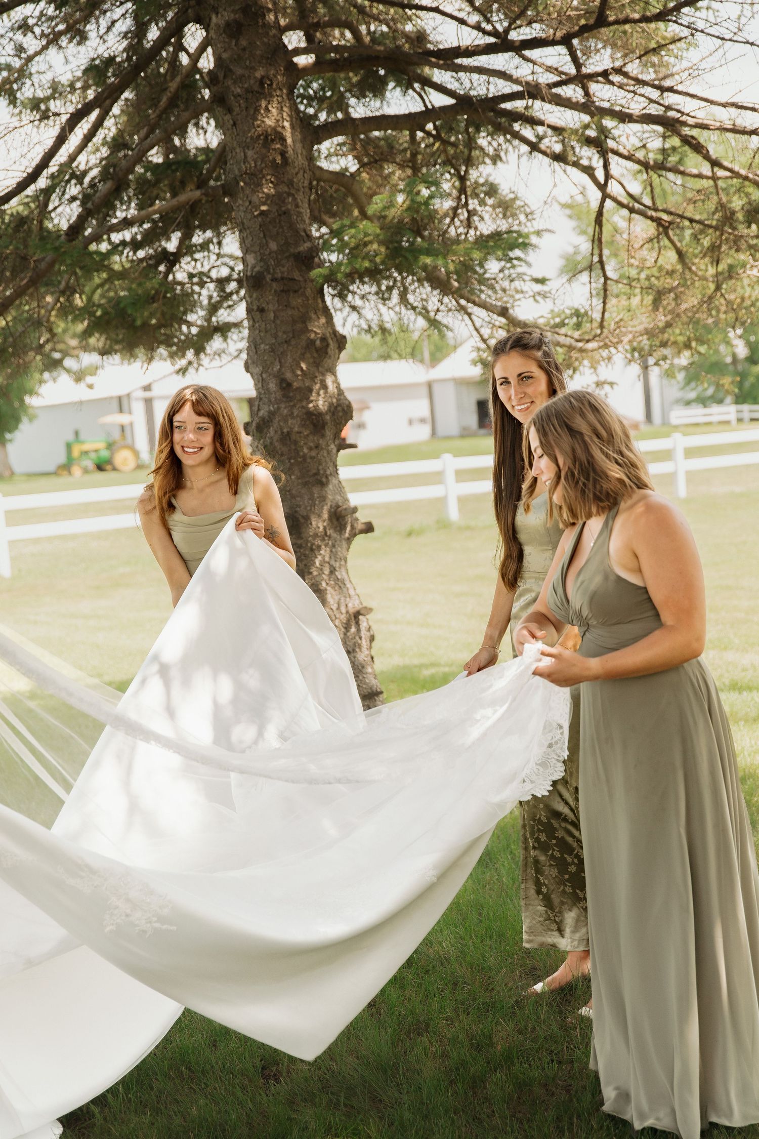 Three bridesmaids in sage green dresses joyfully fluffing bride's white wedding gown train under a tree.