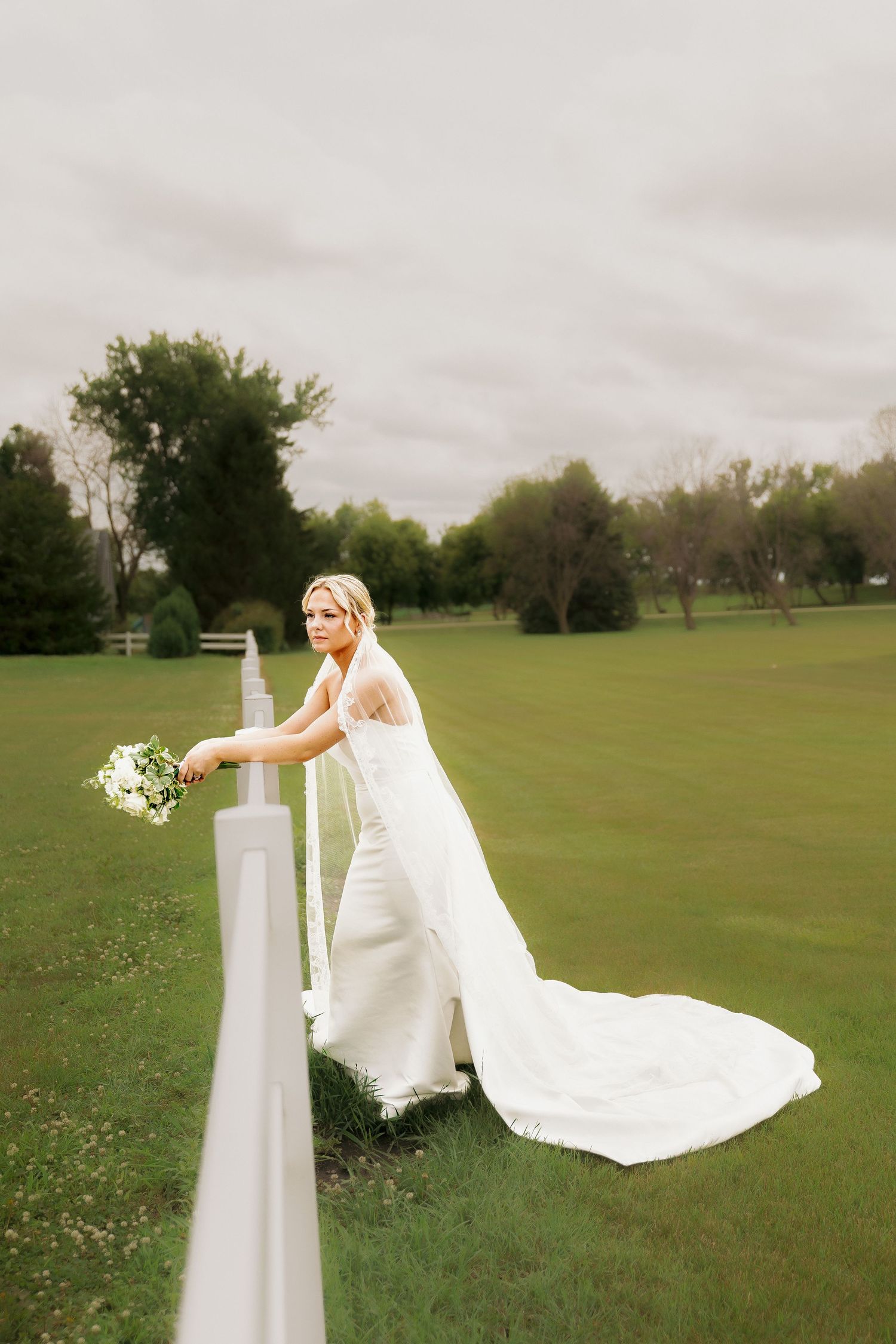 Elegant bride in white gown with long lace veil holds bouquet while leaning on white fence at outdoor wedding.