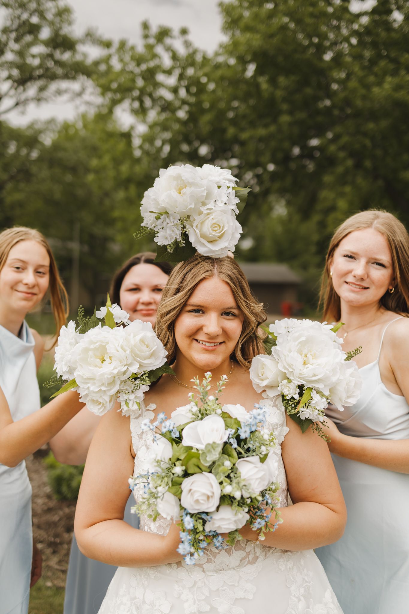 Smiling bride in white lace gown holds bouquet while bridesmaids in blue dresses display white floral arrangements.