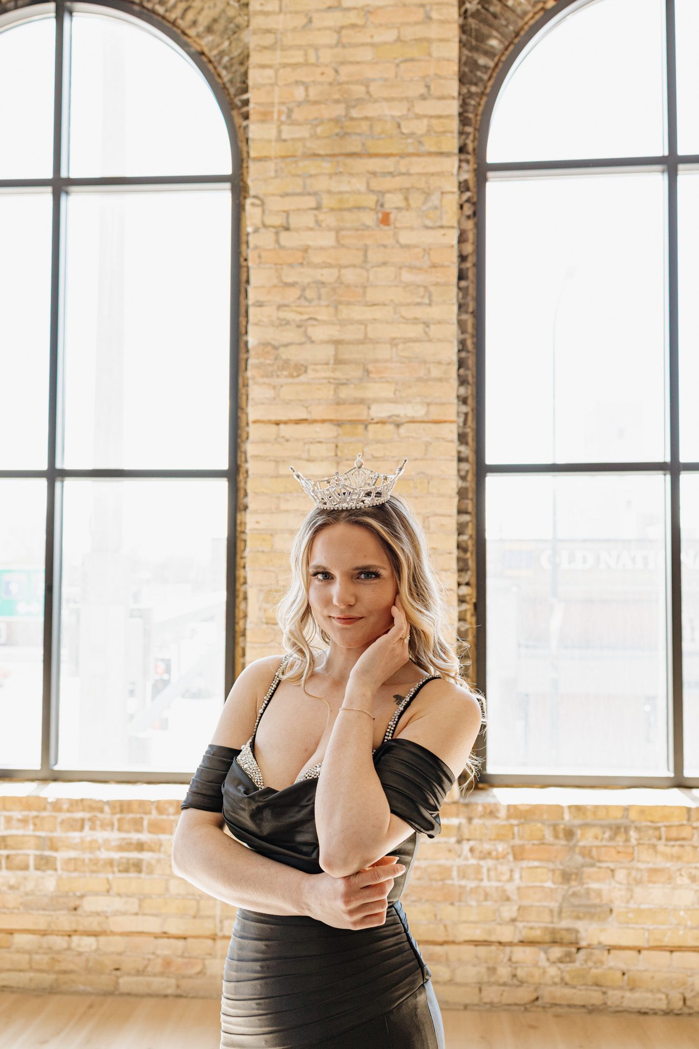 Blonde woman wearing a crystal crown and black off-shoulder dress poses in a loft with brick walls and arched windows.
