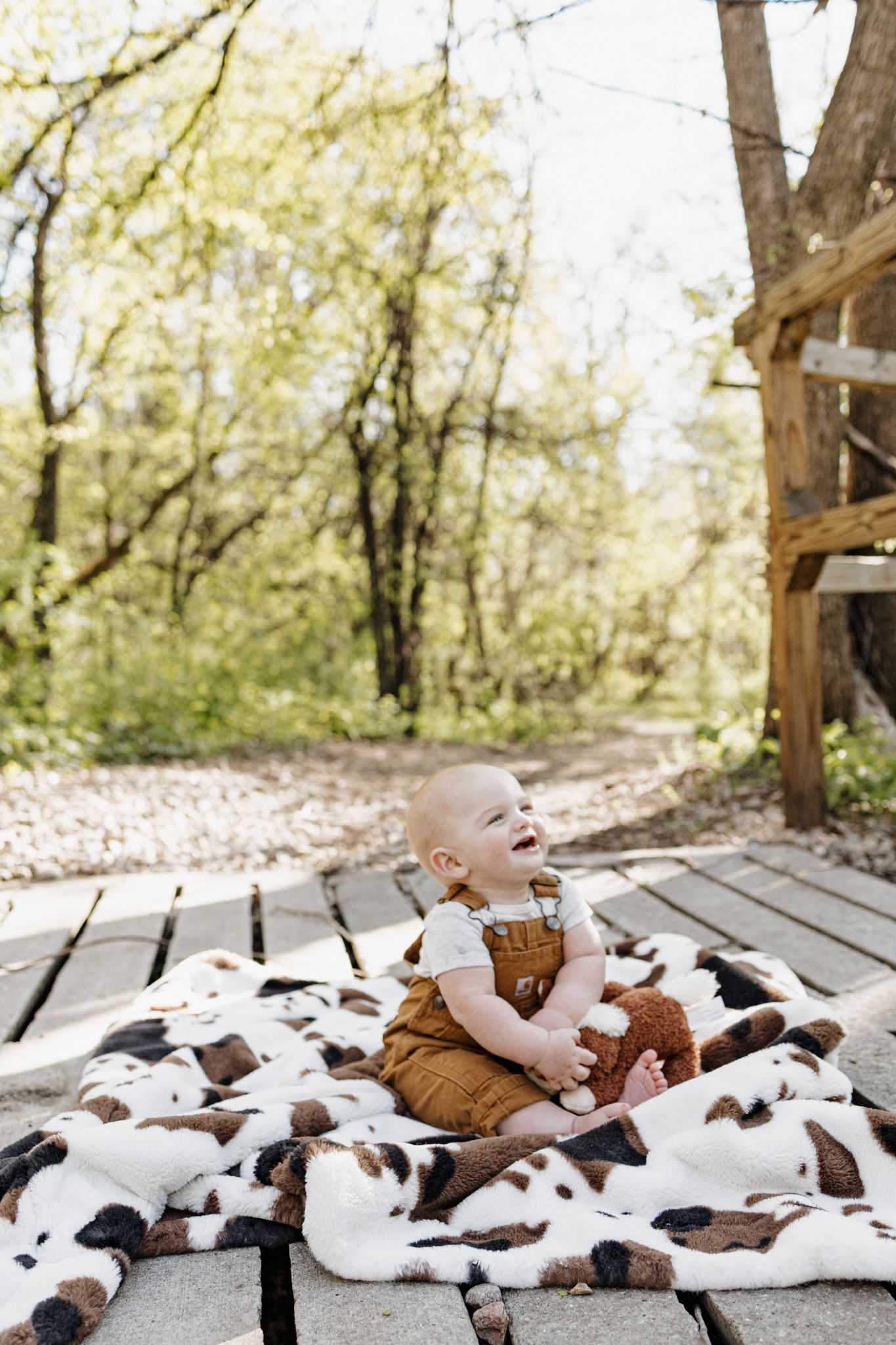 Happy baby in brown overalls sitting on a cowhide blanket on wooden deck, holding a stuffed animal outdoors.