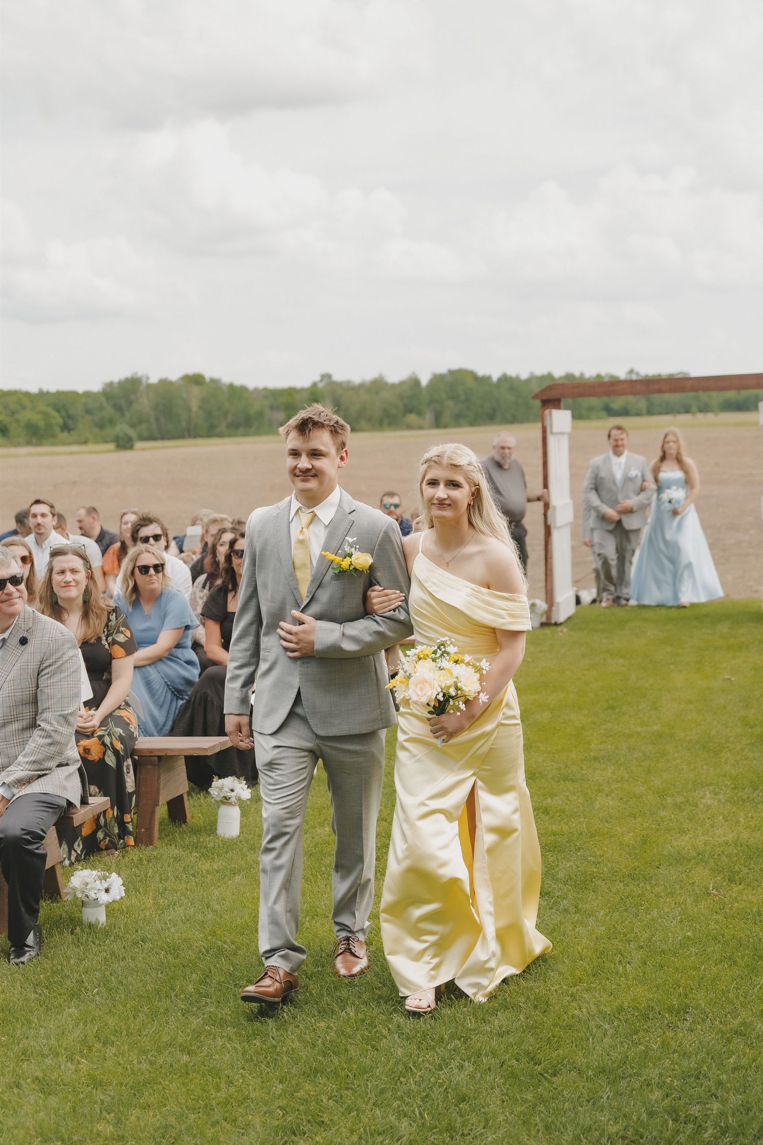 Bridesmaid in yellow off-shoulder gown walks down outdoor aisle with groomsman in gray suit.