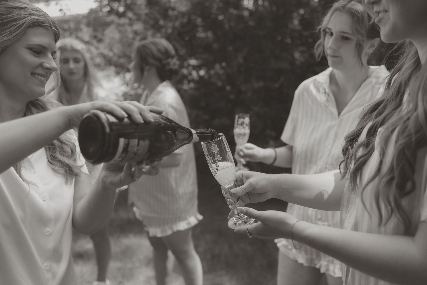 Bridesmaids in white robes toasting with champagne glasses outdoors in a black and white wedding photo.
