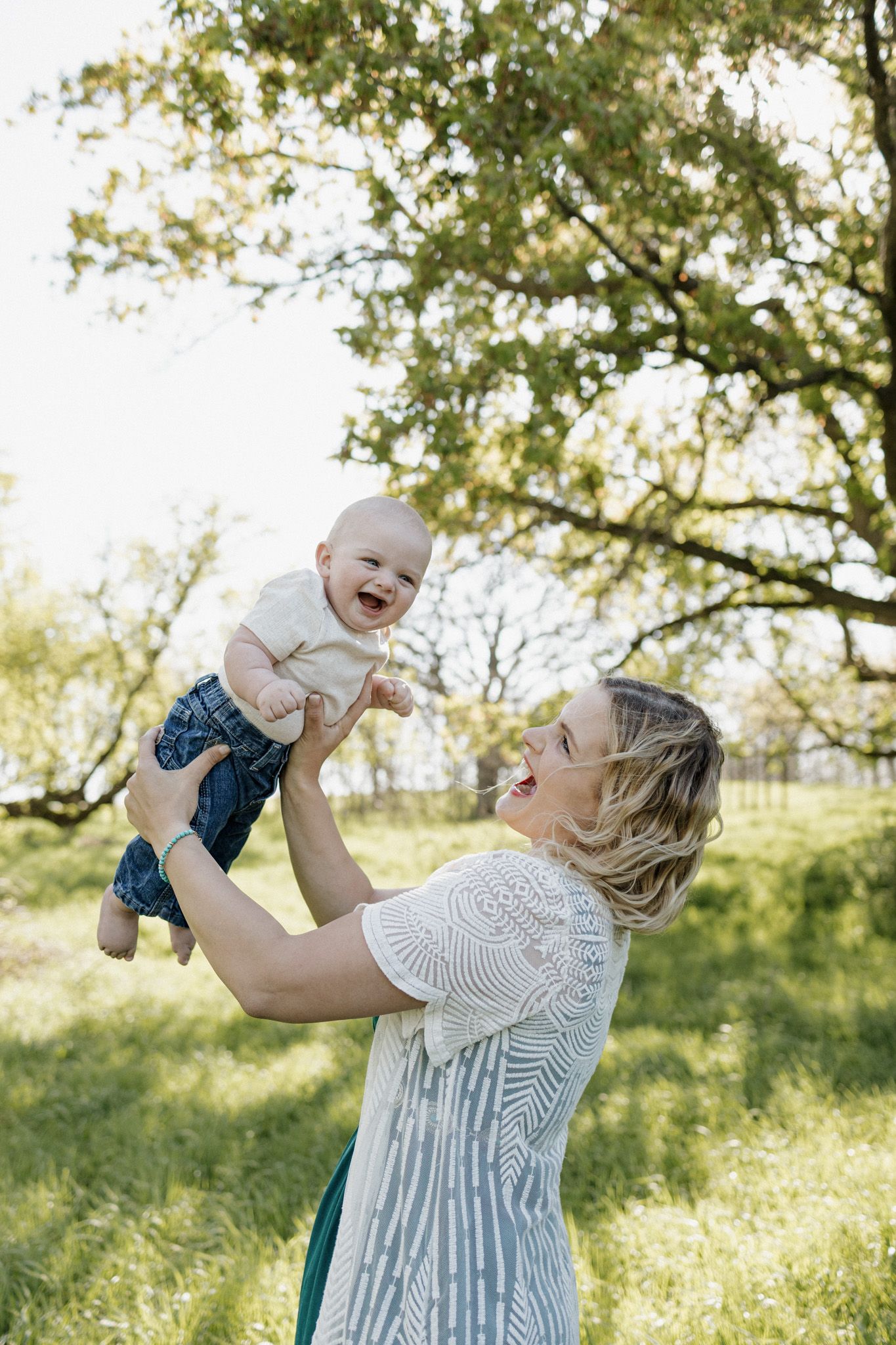 A joyful mother lifts her laughing baby in the air outdoors, surrounded by lush green trees on a sunny day.