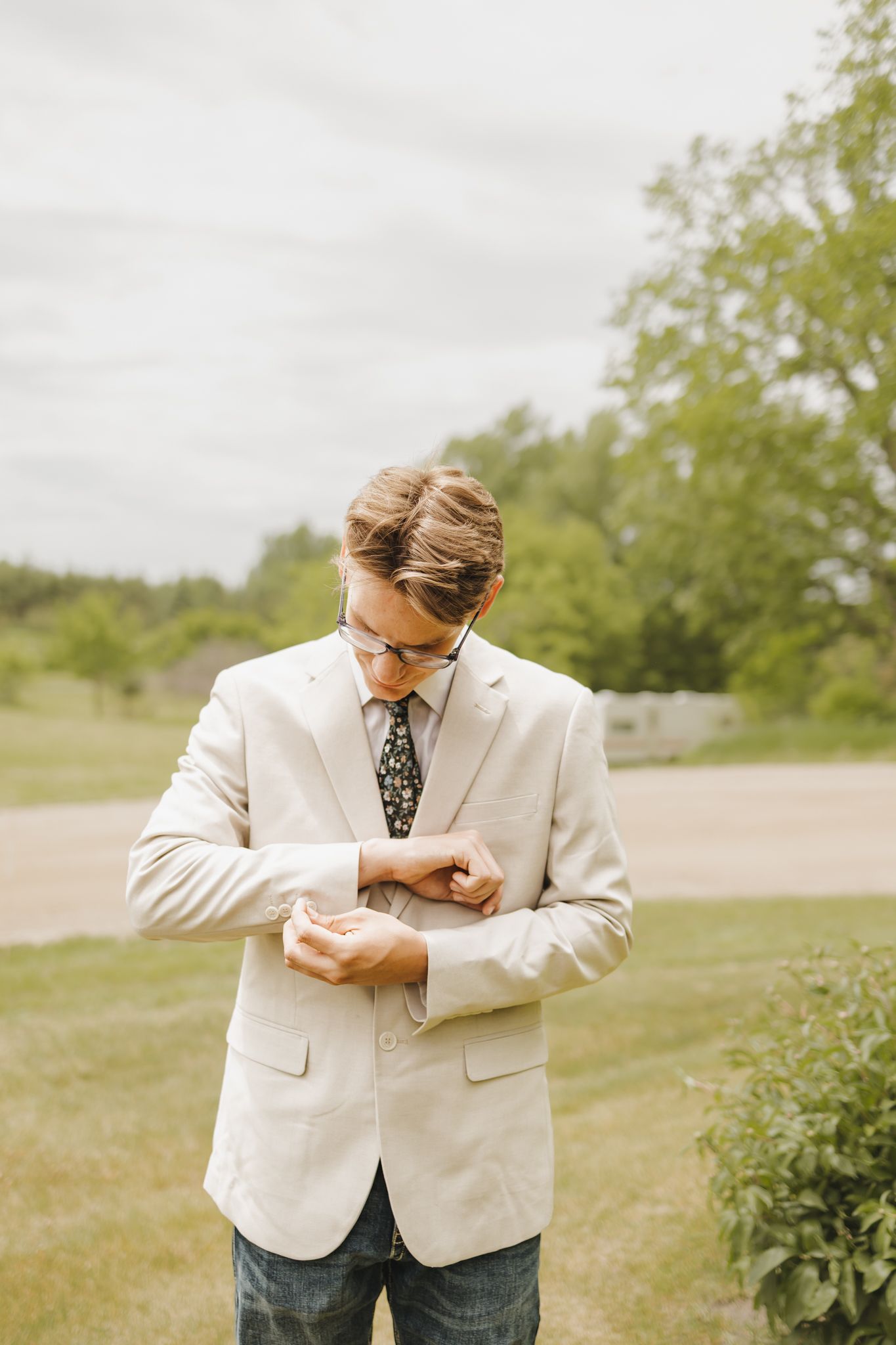 Dapper groom in cream blazer and floral tie adjusting his cufflinks outdoors before wedding ceremony.