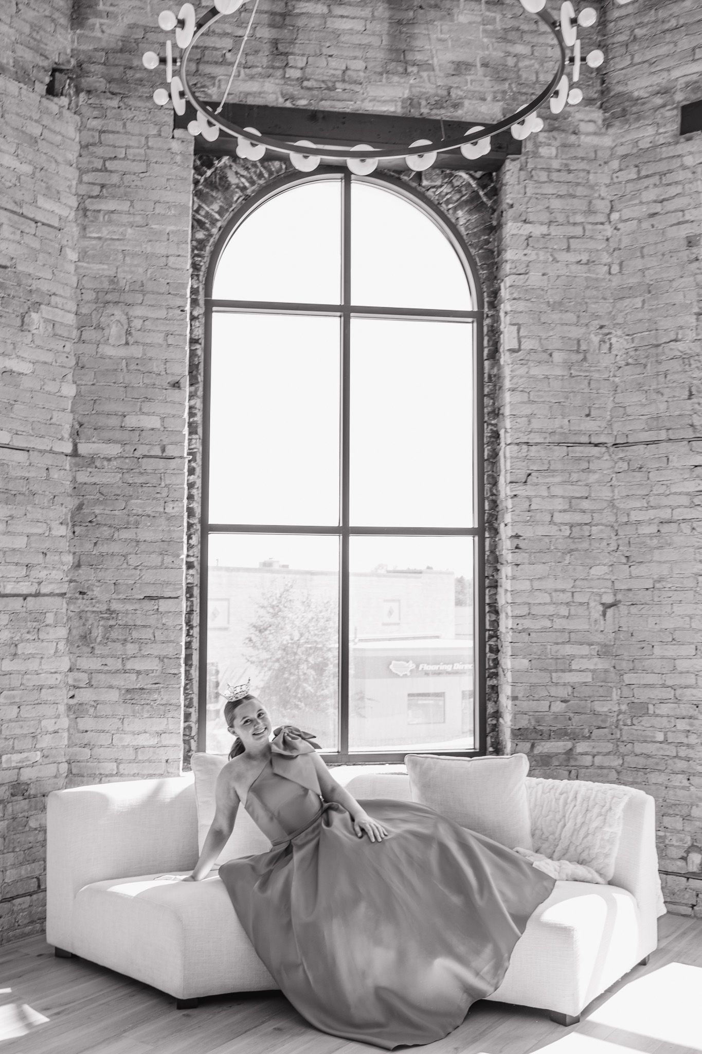 Black and white photo of woman in ball gown on white sofa beneath arched loft window.