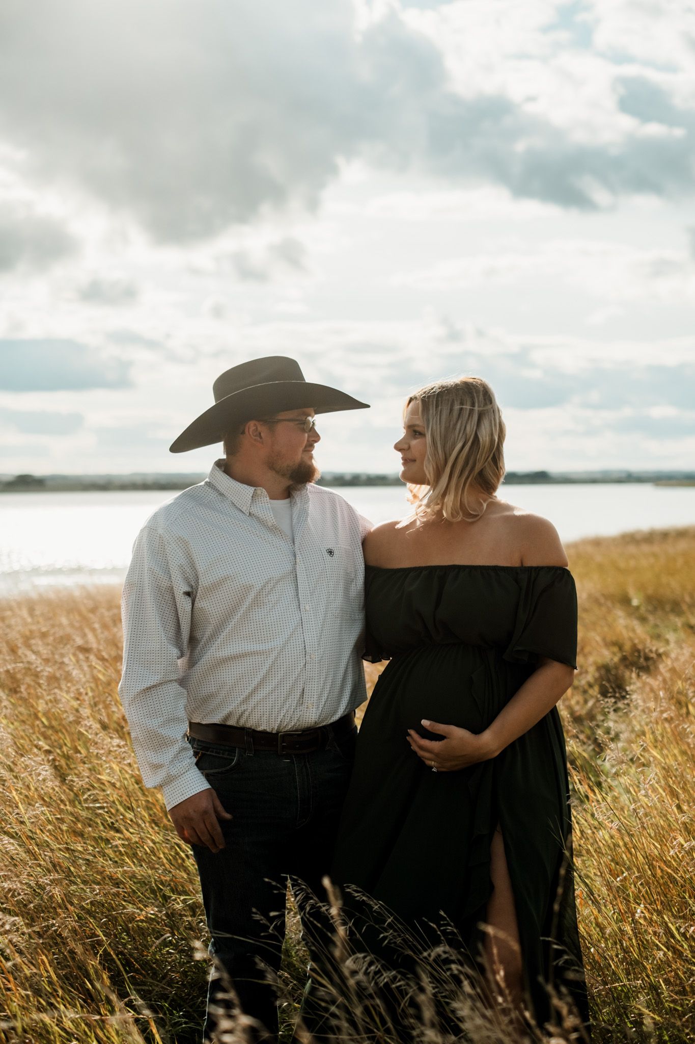 Couple in western attire poses together in tall prairie grass at sunset.
