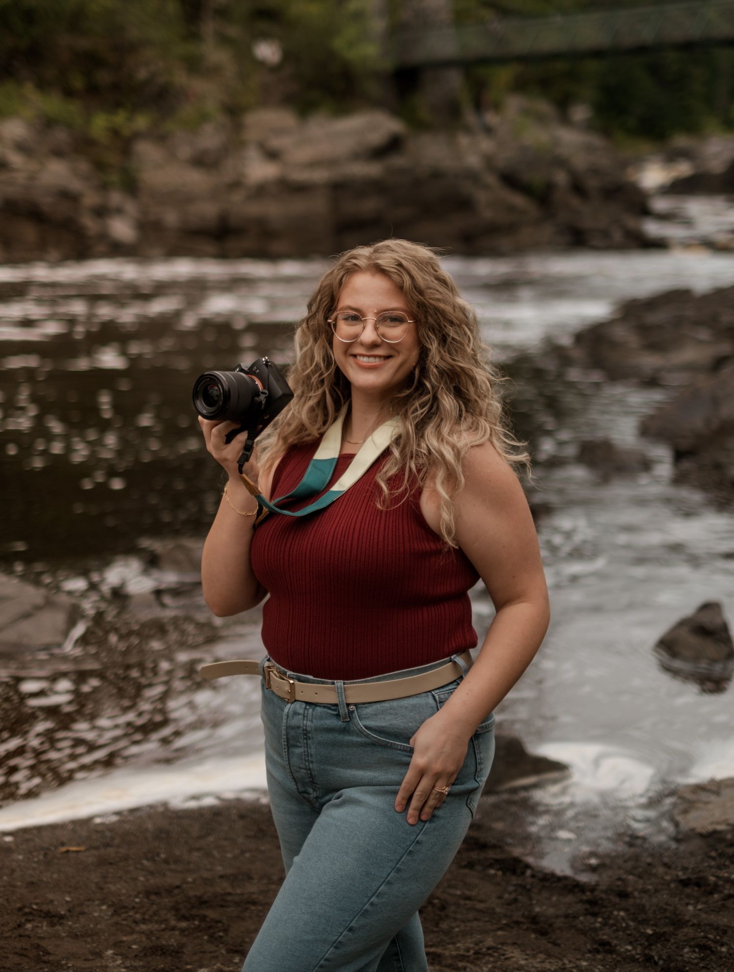 Woman in red top and jeans poses playfully by rocky waterfront.