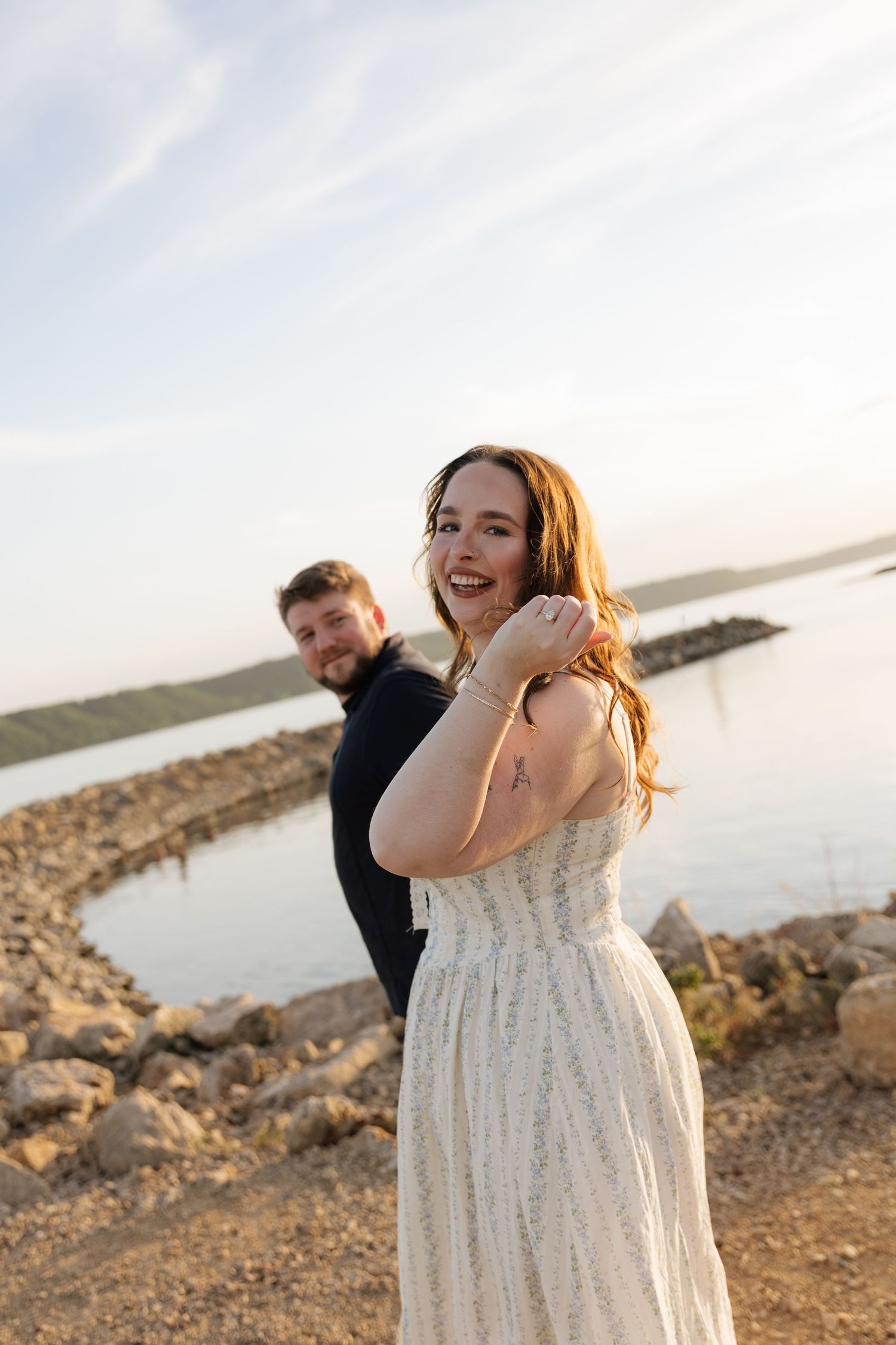 A smiling woman in a white dress poses with a man near a rocky shoreline at sunset.