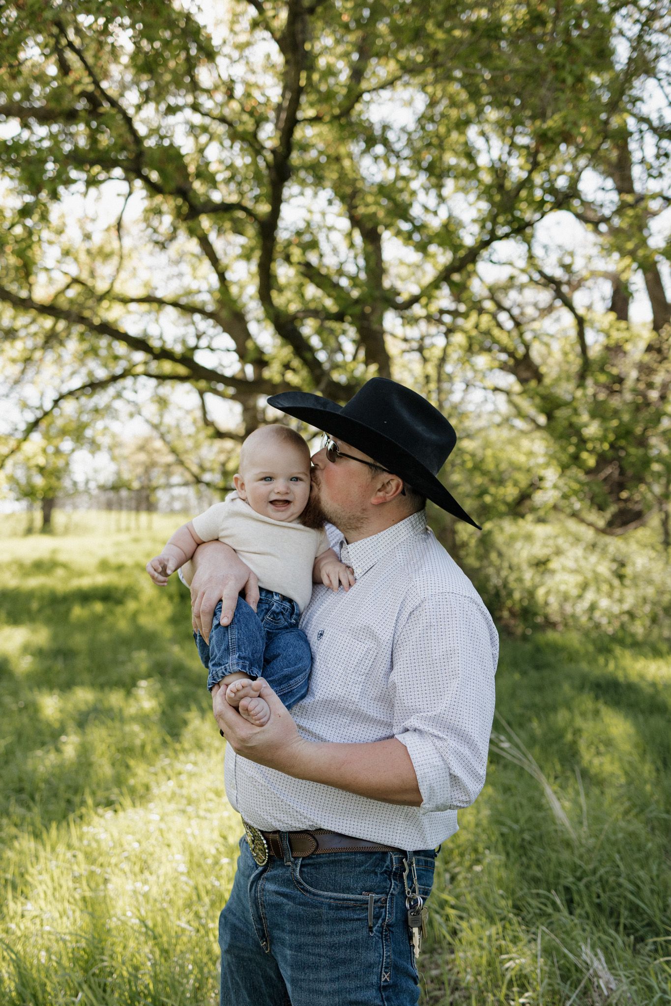 A man in a cowboy hat kisses a smiling baby while standing in a lush green field under a large tree.