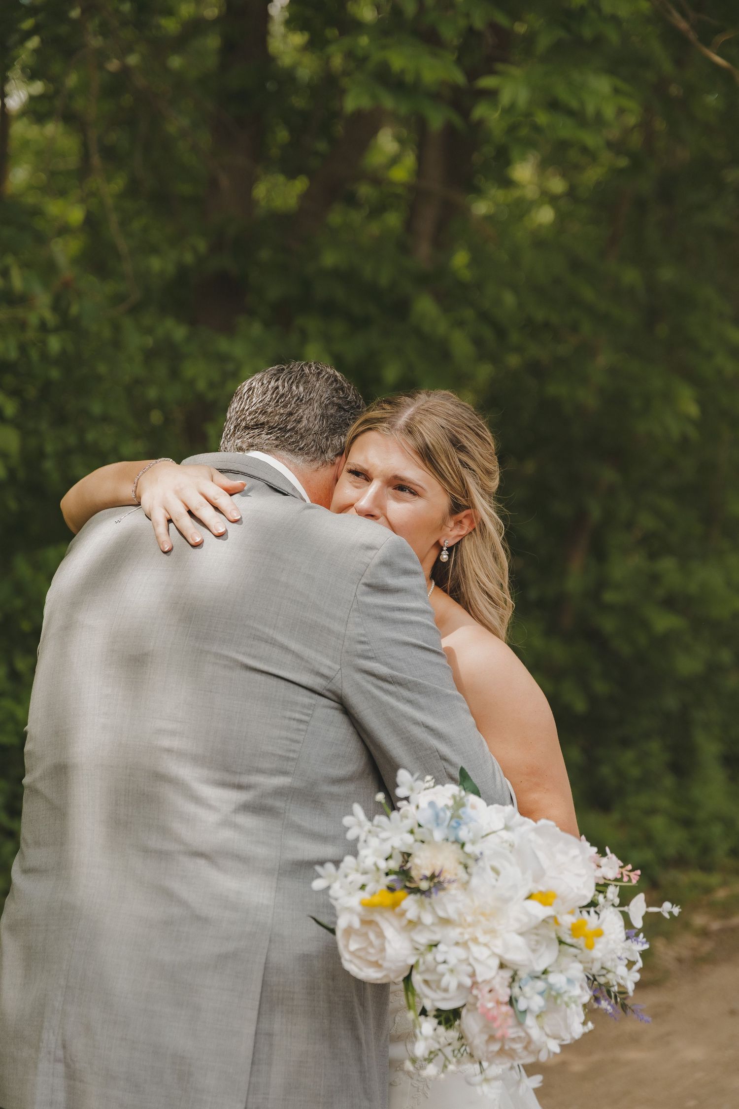Bride hugging a man in a gray suit while holding a white floral bouquet, with lush green trees in the background.