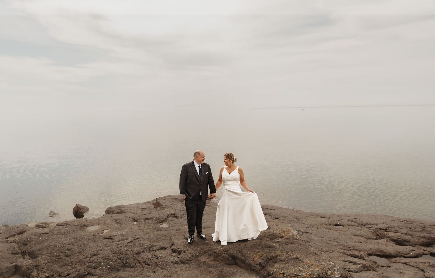Bride and groom holding hands on rocky shoreline with calm misty water backdrop on their wedding day.