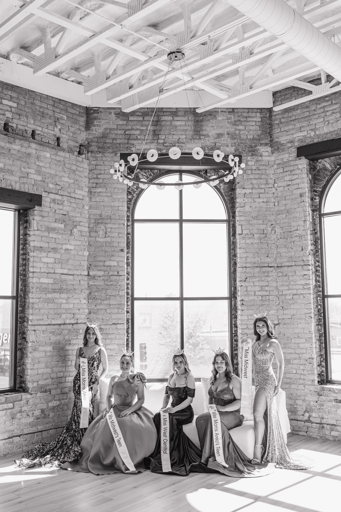 Five pageant contestants in formal gowns and sashes pose in sunlit industrial loft with brick walls.