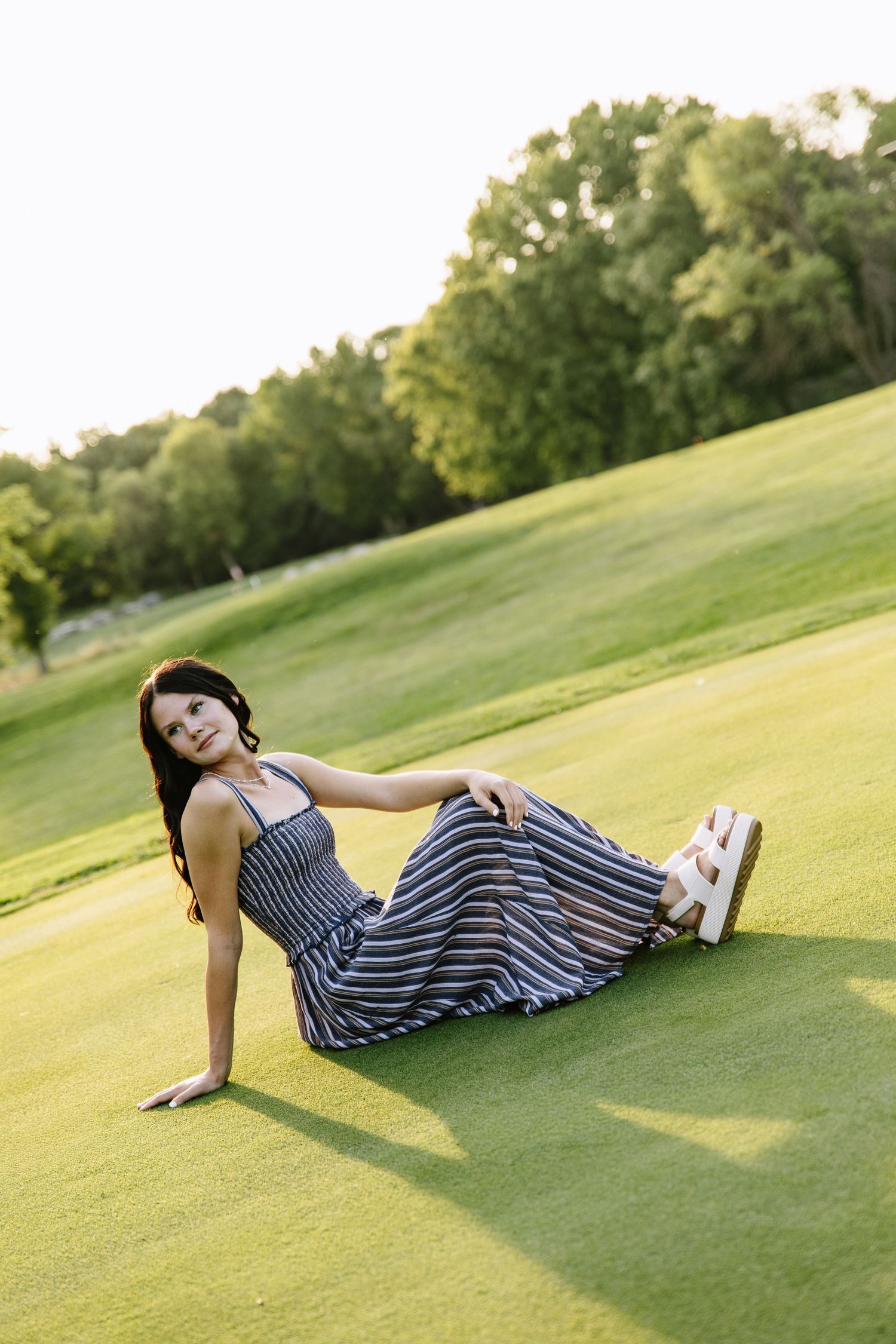 Woman in striped maxi dress sitting on lush green grass in a park, leaning back with trees in background.