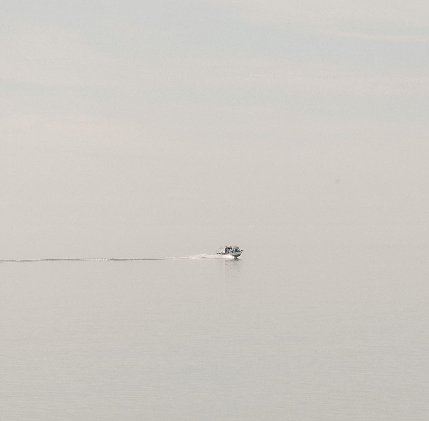 A small boat speeding across a calm, misty gray lake leaving a thin wake trail in the water.