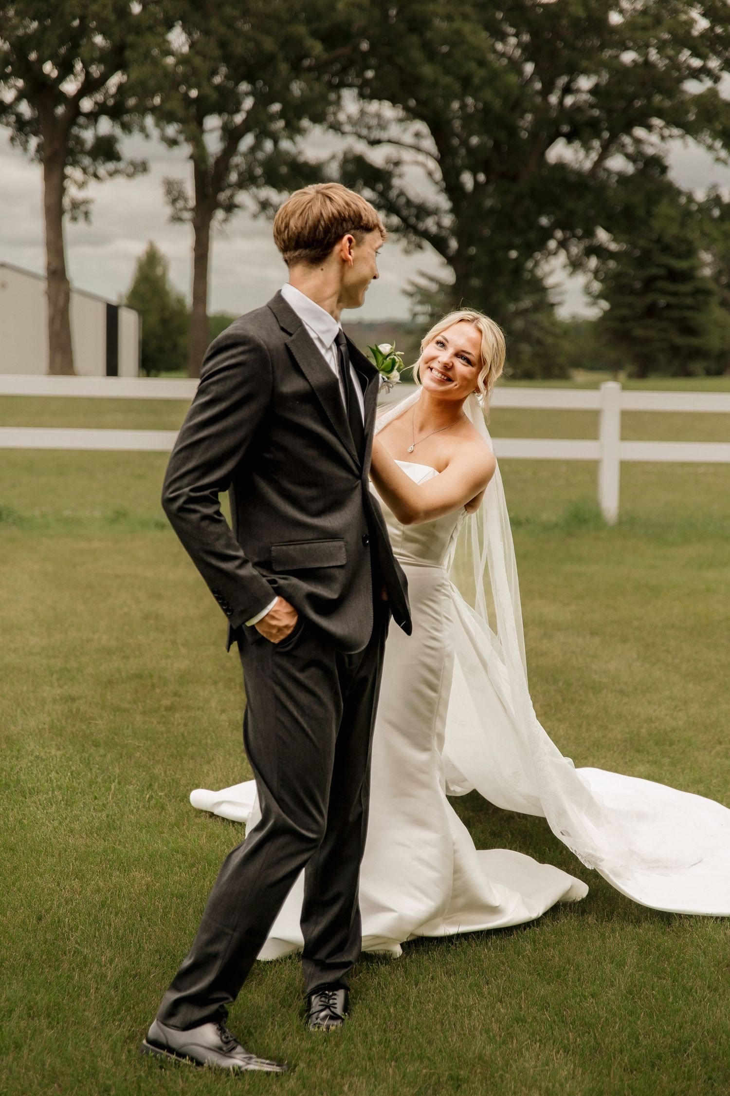 Bride in elegant white mermaid gown and groom in black suit sharing a joyful moment on lush green lawn.