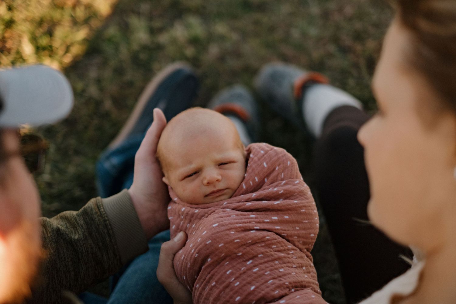 A newborn baby wrapped in a pink swaddle is held during a golden hour outdoor photoshoot.