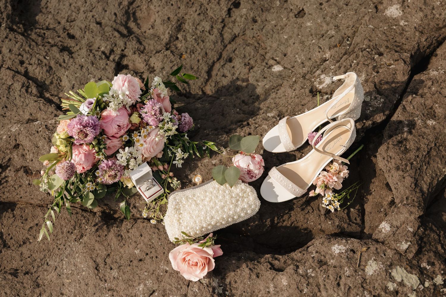 Bridal flat lay with pink floral bouquet, pearl clutch purse, and white strappy heels on rocky surface.