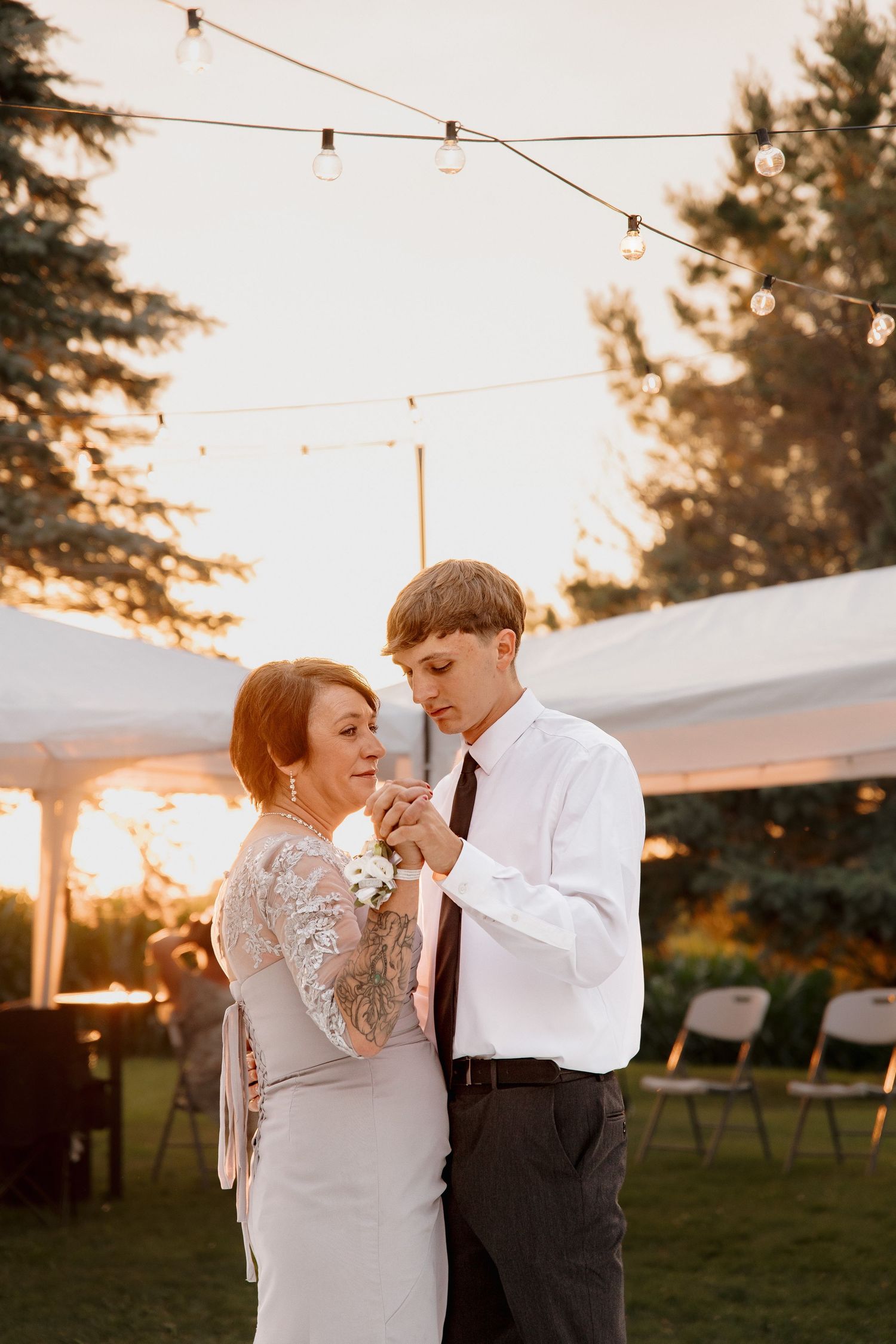 Groom shares a tender mother-son dance under string lights at outdoor wedding reception at golden hour.