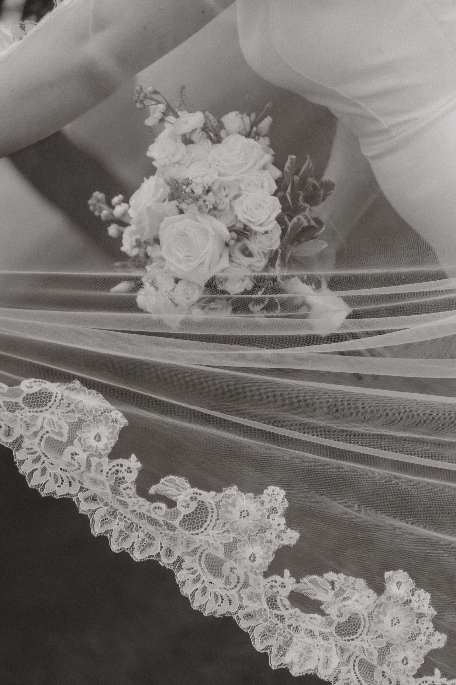 Bride holding a white rose bouquet with elegant lace-trimmed wedding veil spread out in black and white photo.