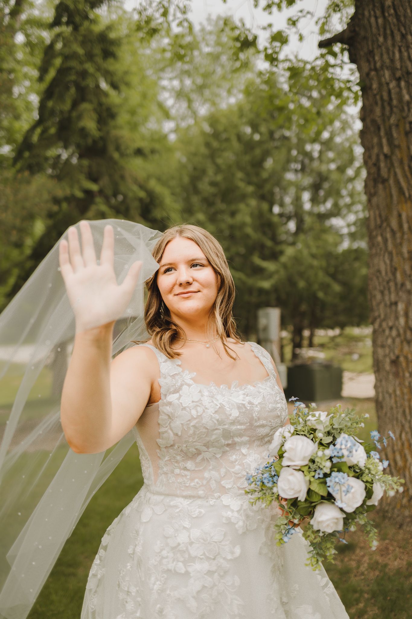 Bride in floral lace wedding gown waves while holding white and blue bouquet with veil flowing near trees.