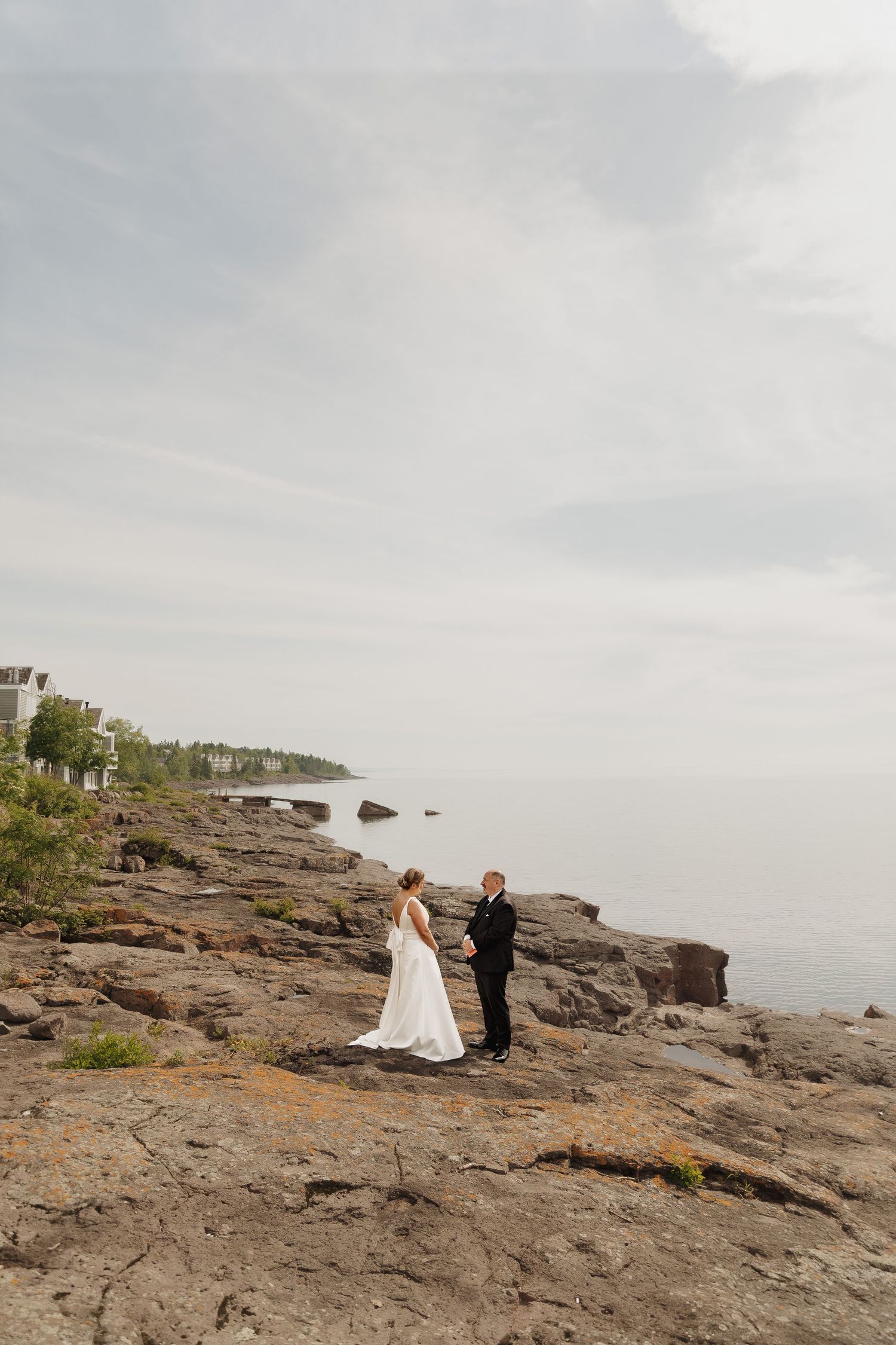 Bride and groom stand on rocky shoreline holding hands with calm ocean and cloudy sky backdrop.