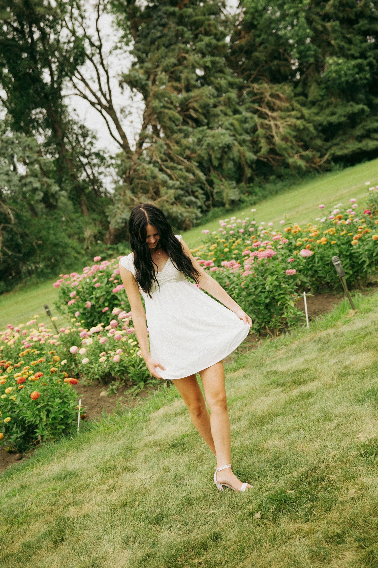 Young woman in white sundress standing on green grass surrounded by colorful flower beds and tall trees in a garden.