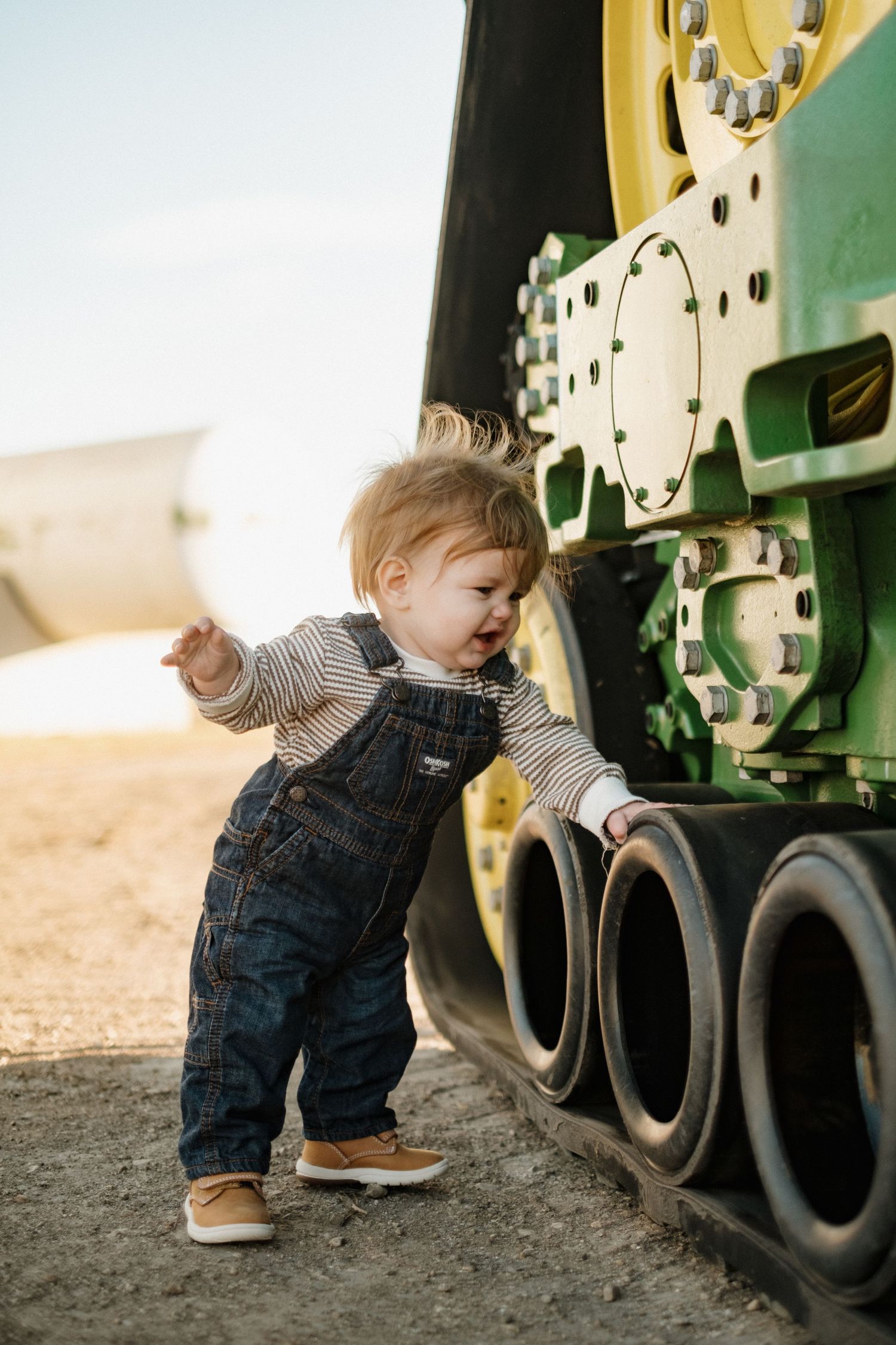 Toddler in denim overalls exploring around large farm equipment wheels.