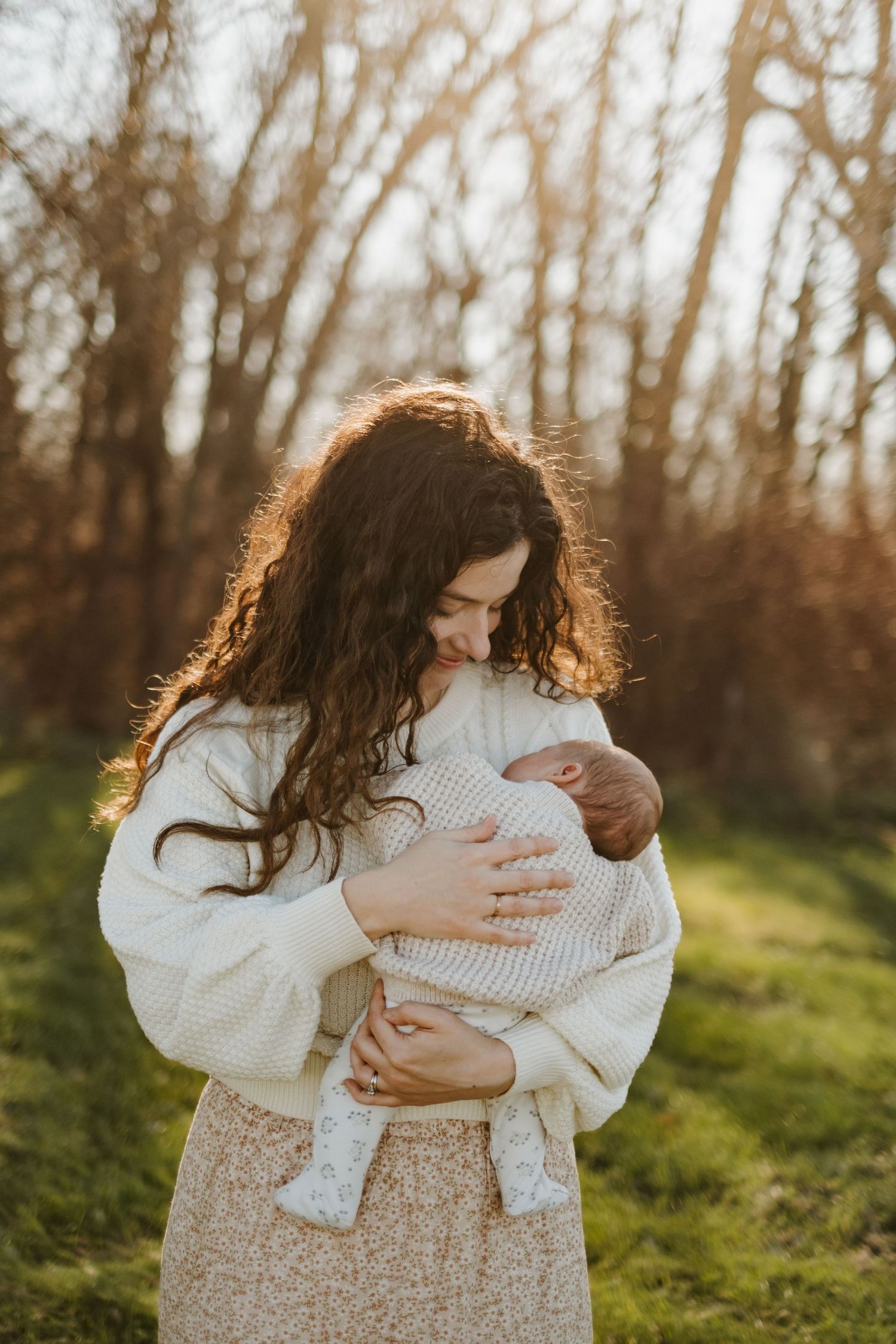 Parent holding infant wrapped in white clothing against sunset-lit forest backdrop.
