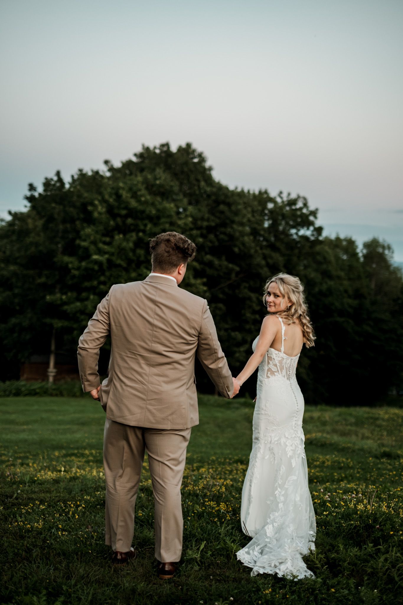 Bride in lace gown and groom in tan suit hold hands walking across green field at dusk.