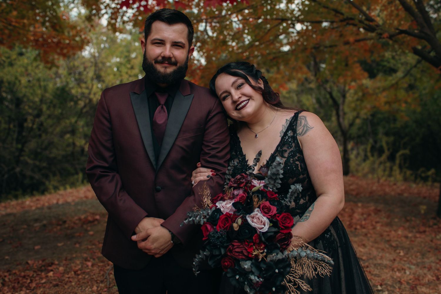 Couple in fall wedding attire, groom in burgundy suit, bride in black gown holding red bouquet, autumn forest backdrop.
