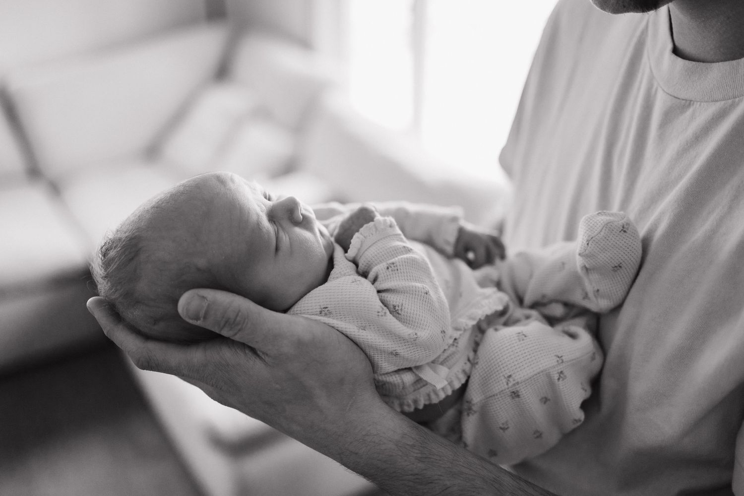 Father cradling a sleeping newborn baby in his hands in a black and white lifestyle photography portrait.