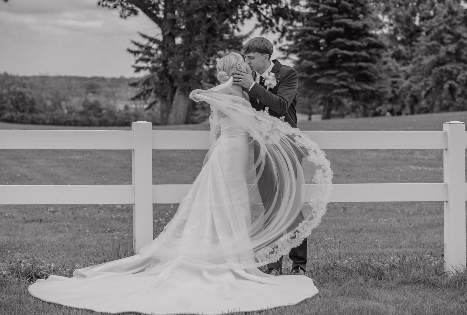 Black and white photo of a bride and groom sharing a kiss, with her veil flowing, beside a white fence.