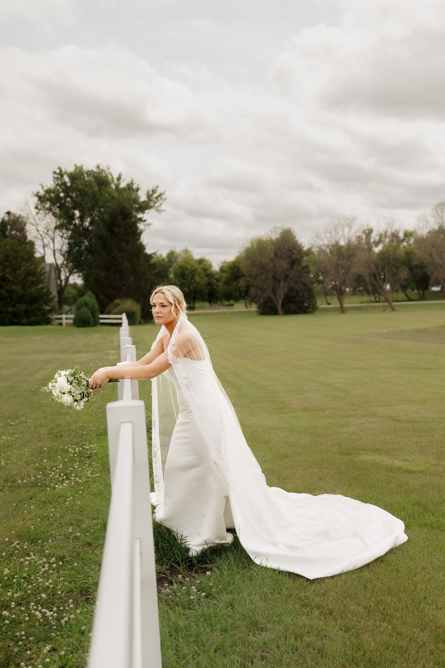 Bride in white gown with train holds bouquet leaning on white fence in lush green outdoor setting.